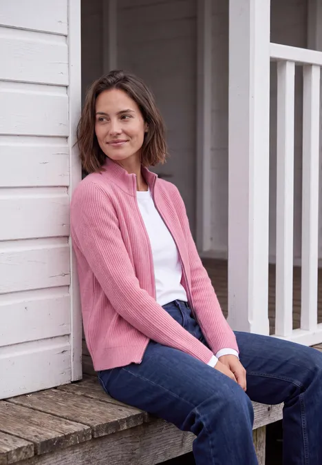 Cardigan mit Stehkragen und Zipper dark blossom melange Frau mit braunen Haaren in rosa Jacke und Jeans sitzt entspannt auf der Veranda eines Holzhauses.