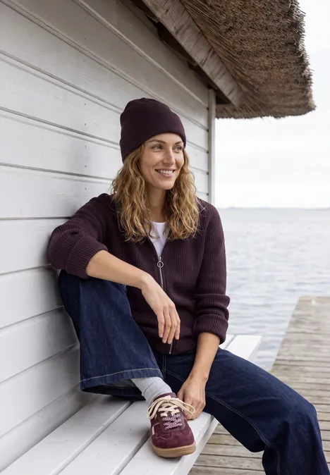 Femme en pull et bonnet violet, appuyée contre un cabanon blanc au bord de l'eau, souriante.