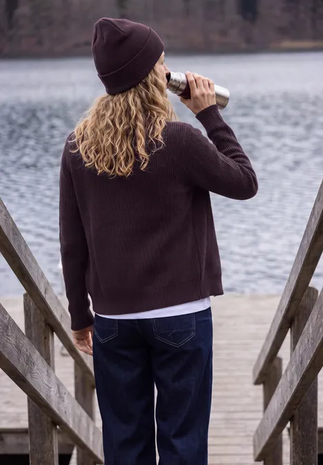 Vue de dos d'une personne en bonnet, pull et jean, buvant au bord d'un lac depuis un ponton.
