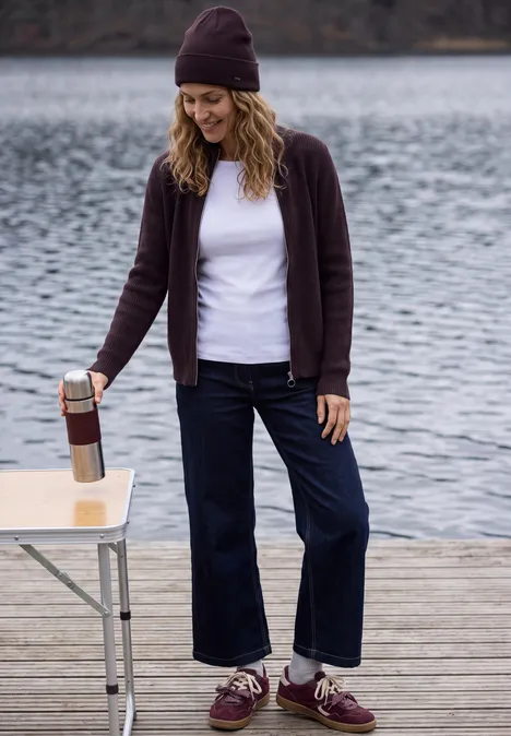 Femme sur un ponton au bord d’un lac, bonnet, t-shirt blanc, gilet, jean large, verse une boisson d’une thermos.