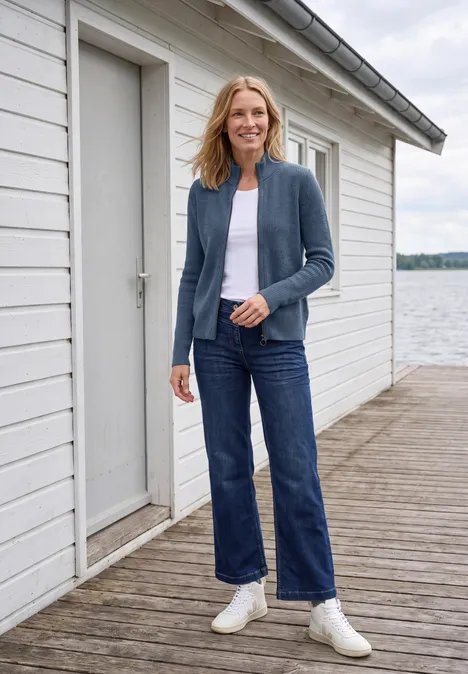 Cardigan mit Stehkragen und Zipper cotton blue melange Frau in Jeans, weißem Shirt und Cardigan steht lächelnd auf einem Steg vor einem weißen Bootshaus am See.