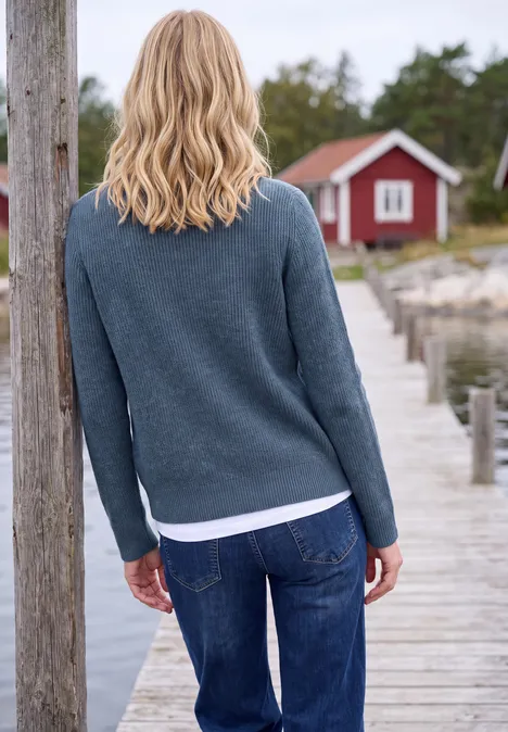 Cardigan mit Stehkragen und Zipper cotton blue melange Eine blonde Frau steht auf einem Holzsteg und blickt auf eine rote Ferienhütte am Wasser.