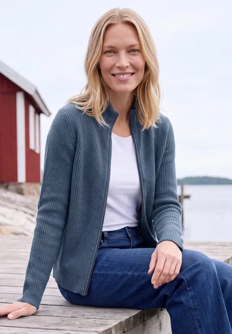 Cardigan mit Stehkragen und Zipper cotton blue melange Eine Frau sitzt lächelnd auf einem Steg vor einem roten Haus am Wasser, trägt Jeans und eine blaue Strickjacke.
