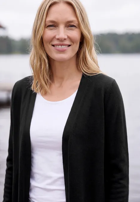 Cardigan long à la coupe ample Black Femme souriante aux cheveux clairs, en t-shirt blanc et gilet sombre devant un lac paisible.