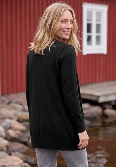 Cardigan long à la coupe ample Black Femme en cardigan noir se tient sur une jetée devant une maison rouge au bord de l'eau et sourit par-dessus son épaule.