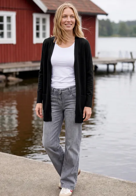 Cardigan long à la coupe ample Black Une femme en jean et gilet se tient sur un ponton devant une maison en bois rouge au bord du lac.