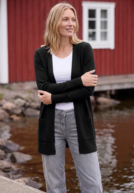 Cardigan long à la coupe ample Black Une femme souriante en gilet noir, top blanc et jean gris pose devant une maison en bois rouge au bord de l'eau.