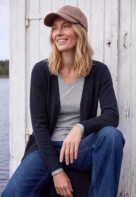 Langer Cardigan in offener Passform urban dark blue Eine lächelnde Frau mit Schirmmütze, grauem Shirt, schwarzer Jacke und Jeans sitzt vor einer Holzwand am Wasser.