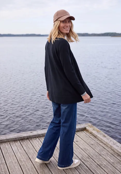 Langer Cardigan in offener Passform urban dark blue Junge Frau mit Kapuzenpulli, Jeans und Mütze steht lächelnd auf einem Holzsteg am See.