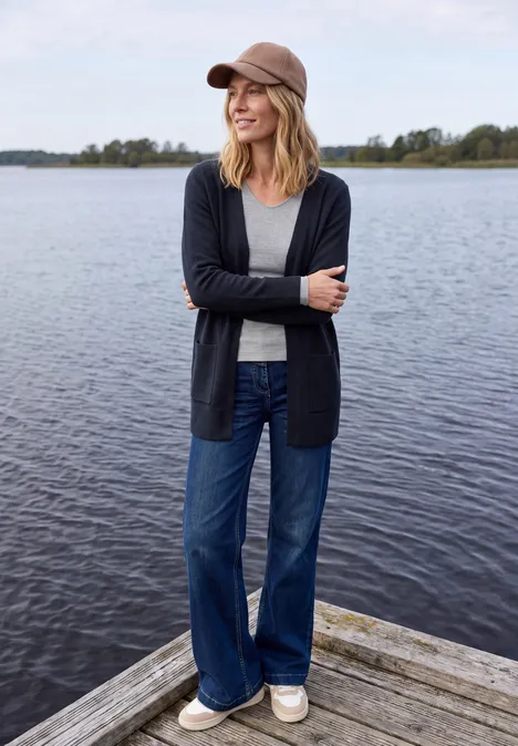 Langer Cardigan in offener Passform urban dark blue Eine lächelnde Frau in Baseballcap, Cardi, Jeans und Sneakers steht auf einem Steg vor ruhigem See.