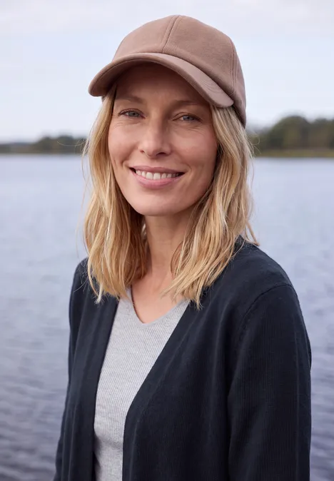 Langer Cardigan in offener Passform urban dark blue Eine blonde Frau mit Kappe lächelt entspannt vor einem See.
