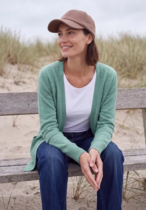 Langer Cardigan in offener Passform jewel green melange Eine Frau mit Mütze und grünem Cardigan sitzt lächelnd auf einer Bank vor Dünen am Strand.