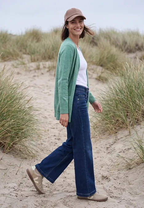 Langer Cardigan in offener Passform jewel green melange Frau in grünem Cardigan, Jeans und Cap steht lächelnd in Dünen mit Strandgras.