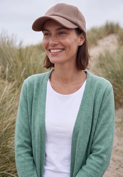 Langer Cardigan in offener Passform jewel green melange Eine lächelnde Frau trägt eine grüne Strickjacke, weißes T-Shirt und eine beige Mütze an einem Dünenstrand.