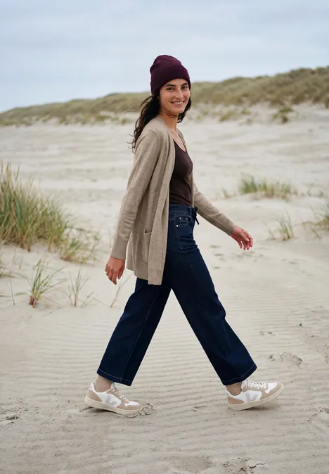 Langer Cardigan in offener Passform toffee cream melange Frau mit Mütze, Cardigan und Jeans spaziert lächelnd am Sandstrand mit Dünen im Hintergrund.