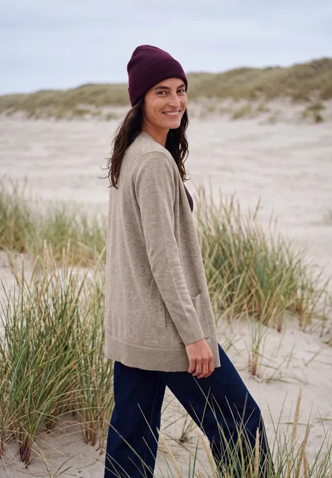 Langer Cardigan in offener Passform toffee cream melange Frau am Strand in beiger Strickjacke, dunkler Mütze und blauer Hose, lächelnd und seitlich zur Kamera gewandt.