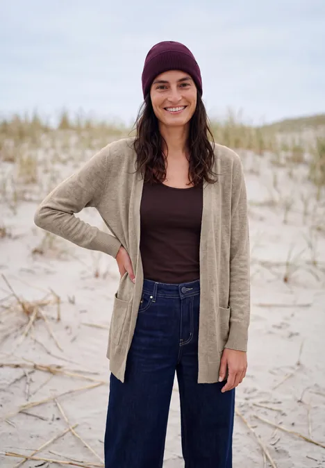 Langer Cardigan in offener Passform toffee cream melange Junge Frau mit Mütze, Cardigan und Jeans lächelt am sandigen Dünenstrand.