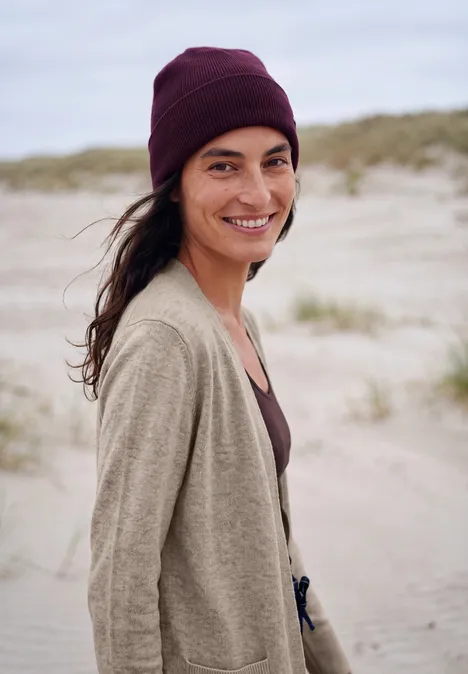 Langer Cardigan in offener Passform toffee cream melange Frau mit dunkelroter Mütze und beige Kleidungsstücken lächelt am windigen Sandstrand vor Dünen.
