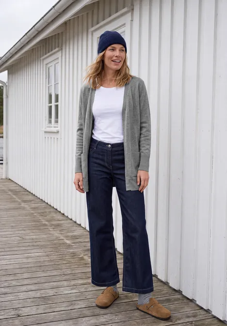 Langer Cardigan in offener Passform sporty grey melange Frau in lässigem Outfit (Beanie, Cardigan, weißes T-Shirt, weite Jeans, Loafers) vor weißem Holzhaus.