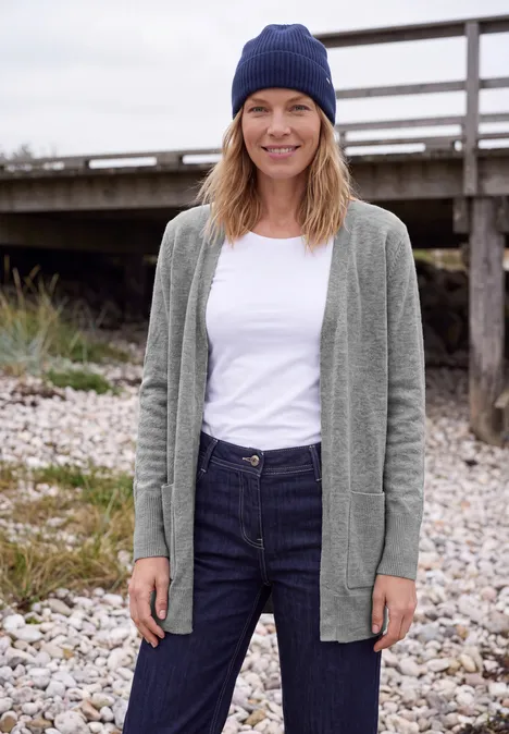 Langer Cardigan in offener Passform sporty grey melange Eine lächelnde Frau im grauen Cardigan, weißem Shirt, Jeans und blauer Mütze steht entspannt am steinigen Ufer vor einem Holzsteg.