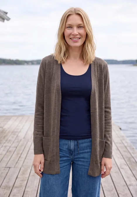 Langer Cardigan in offener Passform mushroom taupe melange Eine lächelnde Frau in Jeans, Top und Cardigan steht auf einem Steg am Wasser.