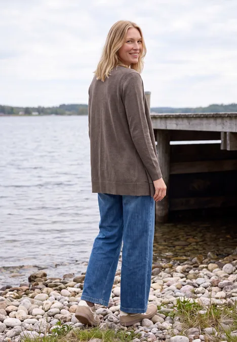 Langer Cardigan in offener Passform mushroom taupe melange Eine Frau in Jeans und Jacke steht lächelnd am Ufer eines Sees, im Hintergrund ein Steg.
