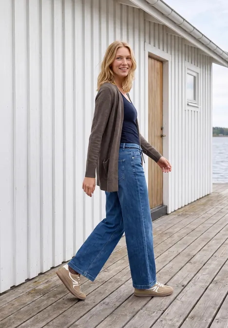 Langer Cardigan in offener Passform mushroom taupe melange Eine lächelnde Frau in Jeans und Strickjacke spaziert auf einer Holzterrasse am Wasser vor einem weißen Haus.