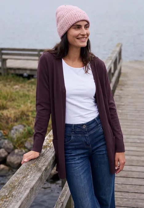 Langer Cardigan in offener Passform eggplant red melange Eine Frau in rosa Mütze und lila Cardigan steht lächelnd auf einem hölzernen Steg am See.