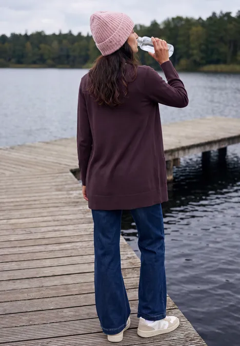 Langer Cardigan in offener Passform eggplant red melange Frau in rosa Mütze und violettem Pullover trinkt am Steg Wasser, Blick zum See und Wald.
