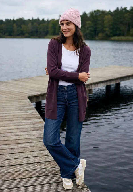 Langer Cardigan in offener Passform eggplant red melange Eine lächelnde Frau in Cardigan, Jeans und Mütze steht auf einem Steg am See.