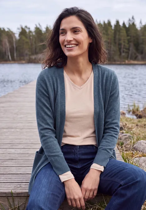 Cardigan long à la coupe ample cotton blue melange Femme souriante assise sur une jetée en bois au bord d’un lac, en cardigan et jean, forêt en arrière-plan.