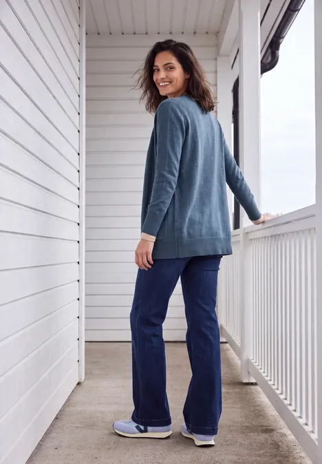 Cardigan long à la coupe ample cotton blue melange Femme en cardigan bleu et jean souriante, regard par-dessus l'épaule sur une terrasse blanche.