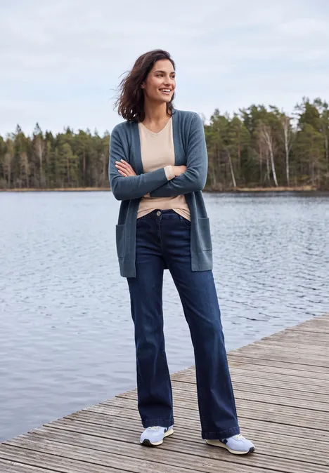 Cardigan long à la coupe ample cotton blue melange Femme décontractée se tient sur une jetée en bois au bord d'un lac, en cardigan, jean et baskets.