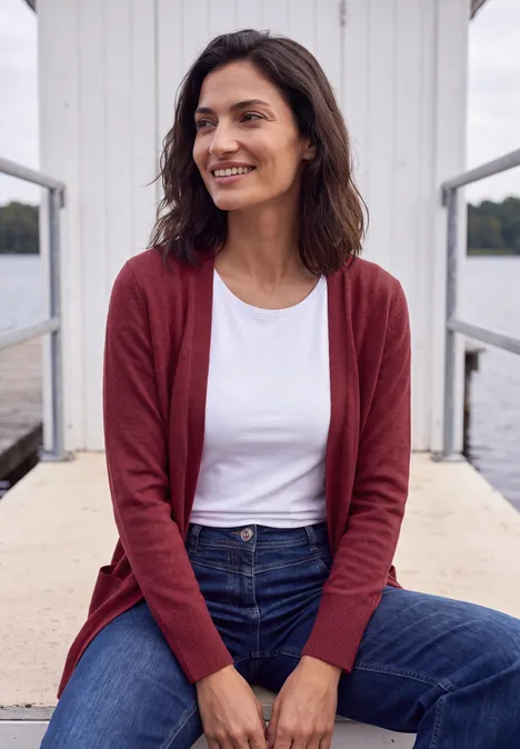 Langer Cardigan in offener Passform rhubarb melange Frau steht entspannt auf einem Steg, trägt roten Cardigan und Jeans.