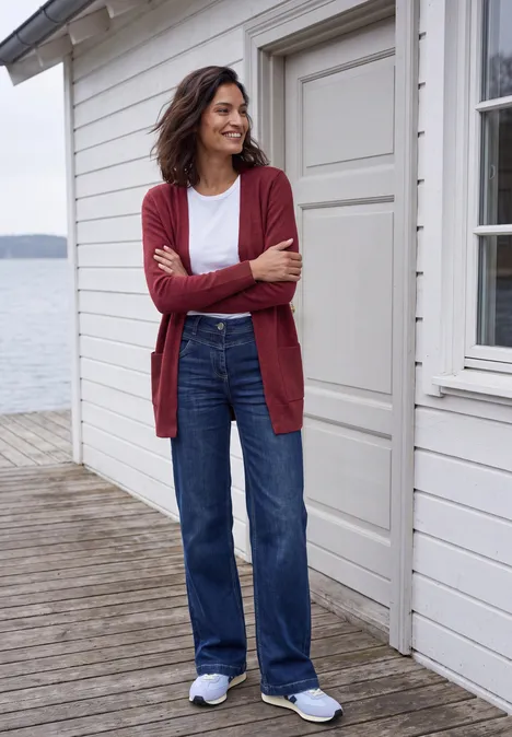 Langer Cardigan in offener Passform rhubarb melange Frau in rotem Cardigan, weißem Shirt und blauen Jeans lehnt lächelnd an Holzwand auf einer Seeterrasse.