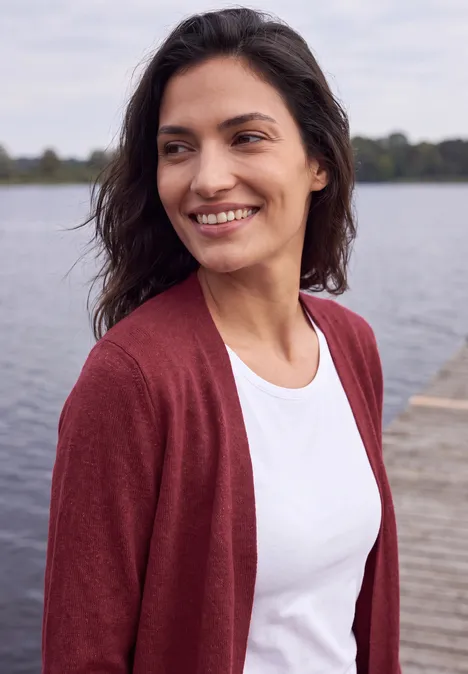 Langer Cardigan in offener Passform rhubarb melange Eine lächelnde Frau im weißen T-Shirt und roter Strickjacke steht an einem Steg am See.