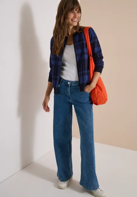 Langarm Cardigan mit Rundhals und Zipper urban dark blue Frau in kariertem Hemd, weißem Shirt, Jeans, weißen Sneakern und orangefarbener Tasche vor neutralem Hintergrund.