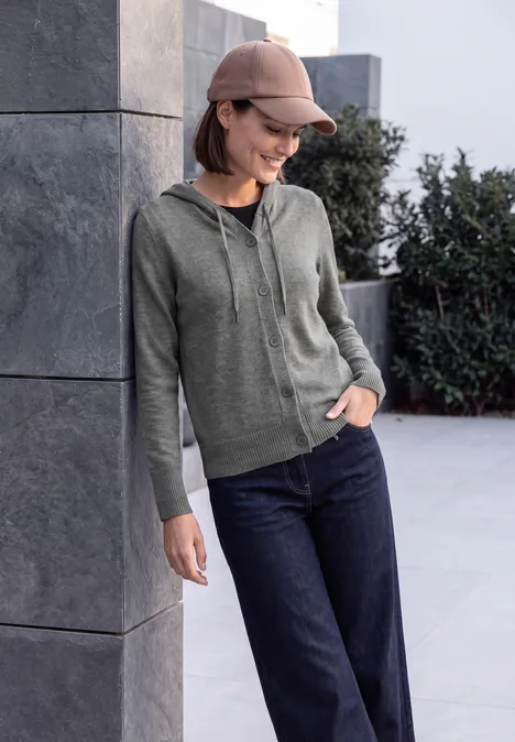 Eine junge Frau in grüner Kapuzenjacke, Jeans und Mütze lehnt lächelnd an einer Betonwand.