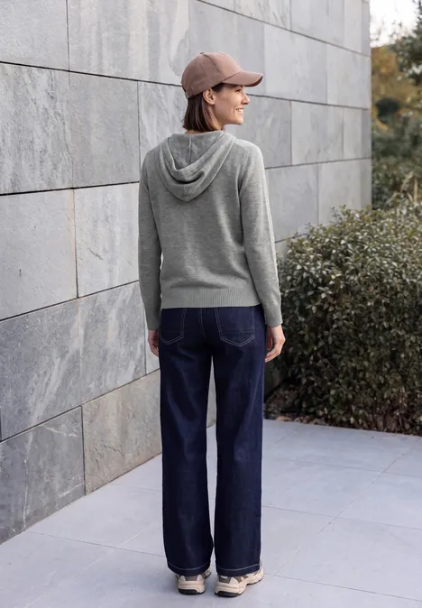 Eine Person mit grauem Kapuzenpullover, weiter Jeans und Kappe steht seitlich vor einer Steinwand.