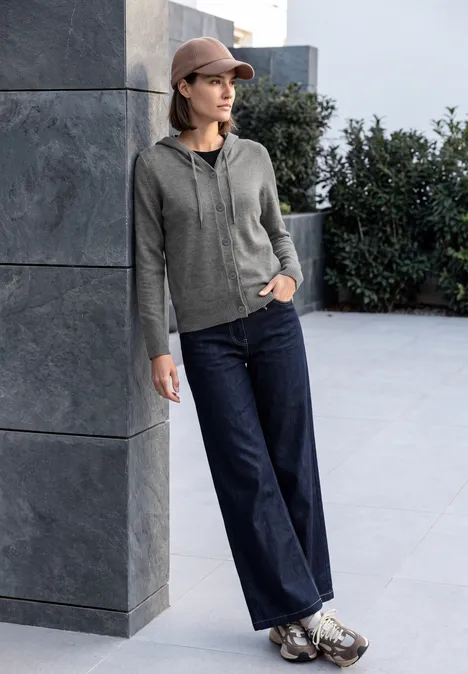 Eine Frau in grauem Kapuzenpullover, Jeans und Cap lehnt draußen lässig an einer Wand.