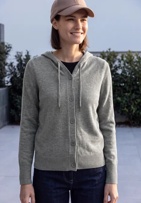 Frau in grauer Strickjacke mit Kapuze, Jeans und Cap, lächelnd im Freien vor Hecke.