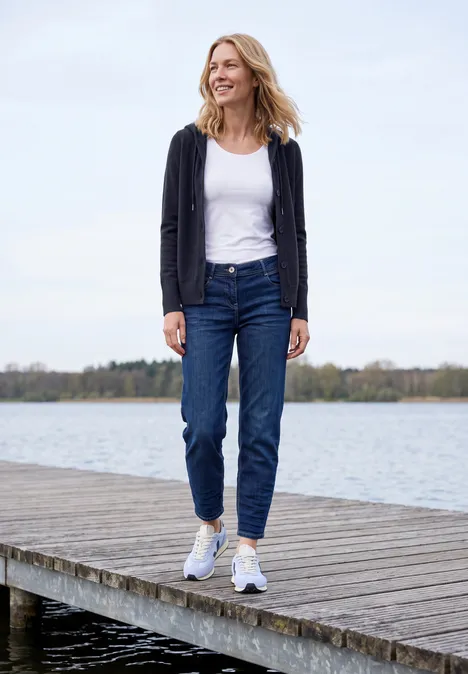 Frau läuft auf einem Steg am See, trägt Jeans, weißes Top und Cardigan