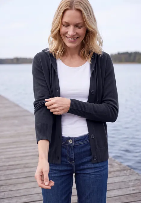 Frau steht lächelnd auf einem Holzsteg am See, trägt Jeans, weißes Shirt und schwarze Strickjacke.