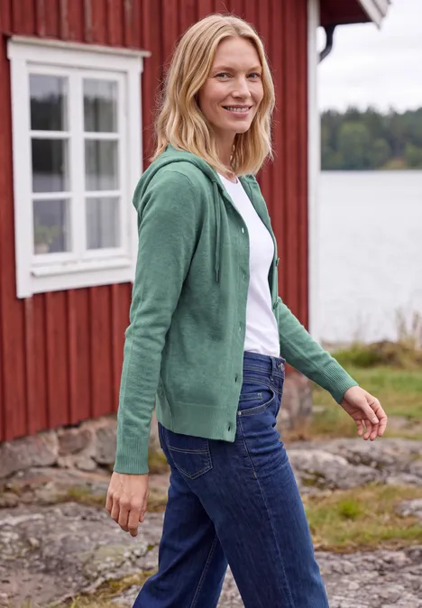Langarm Cardigan mit Kapuze jewel green melange Eine blonde Frau in grünem Kapuzenpullover und Jeans steht lächelnd vor einem roten Holzhaus am See.