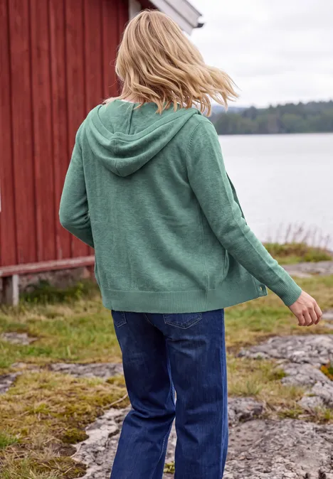 Langarm Cardigan mit Kapuze jewel green melange Eine Person in grüner Kapuzen-Strickjacke und Jeans steht am Ufer vor einem roten Haus, Blick aufs Wasser.