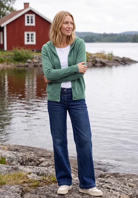 Langarm Cardigan mit Kapuze jewel green melange Frau steht auf Felsen am See, trägt grünen Cardigan, weißes Shirt und Jeans, rotes Haus im Hintergrund.