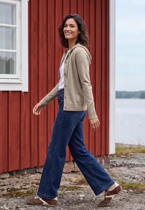 Langarm Cardigan mit Kapuze toffee cream melange Frau in Jeans und Jacke spaziert fröhlich an einem roten Holzhaus am Wasser.