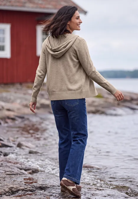 Langarm Cardigan mit Kapuze toffee cream melange Frau im Kapuzenpullover spaziert an einem Seeufer, Blick aufs Wasser, rote Holzhütte im Hintergrund.