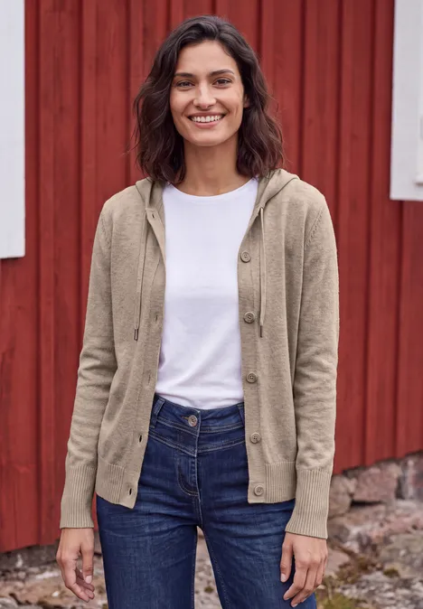 Langarm Cardigan mit Kapuze toffee cream melange Eine lächelnde Frau in Jeans, weißem T-Shirt und beiger Strickjacke vor einer roten Holzwand.
