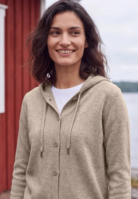 Langarm Cardigan mit Kapuze toffee cream melange Eine lächelnde Frau in beige Kapuzenjacke und weißem Shirt steht draußen vor einem roten Holzhaus am Wasser.